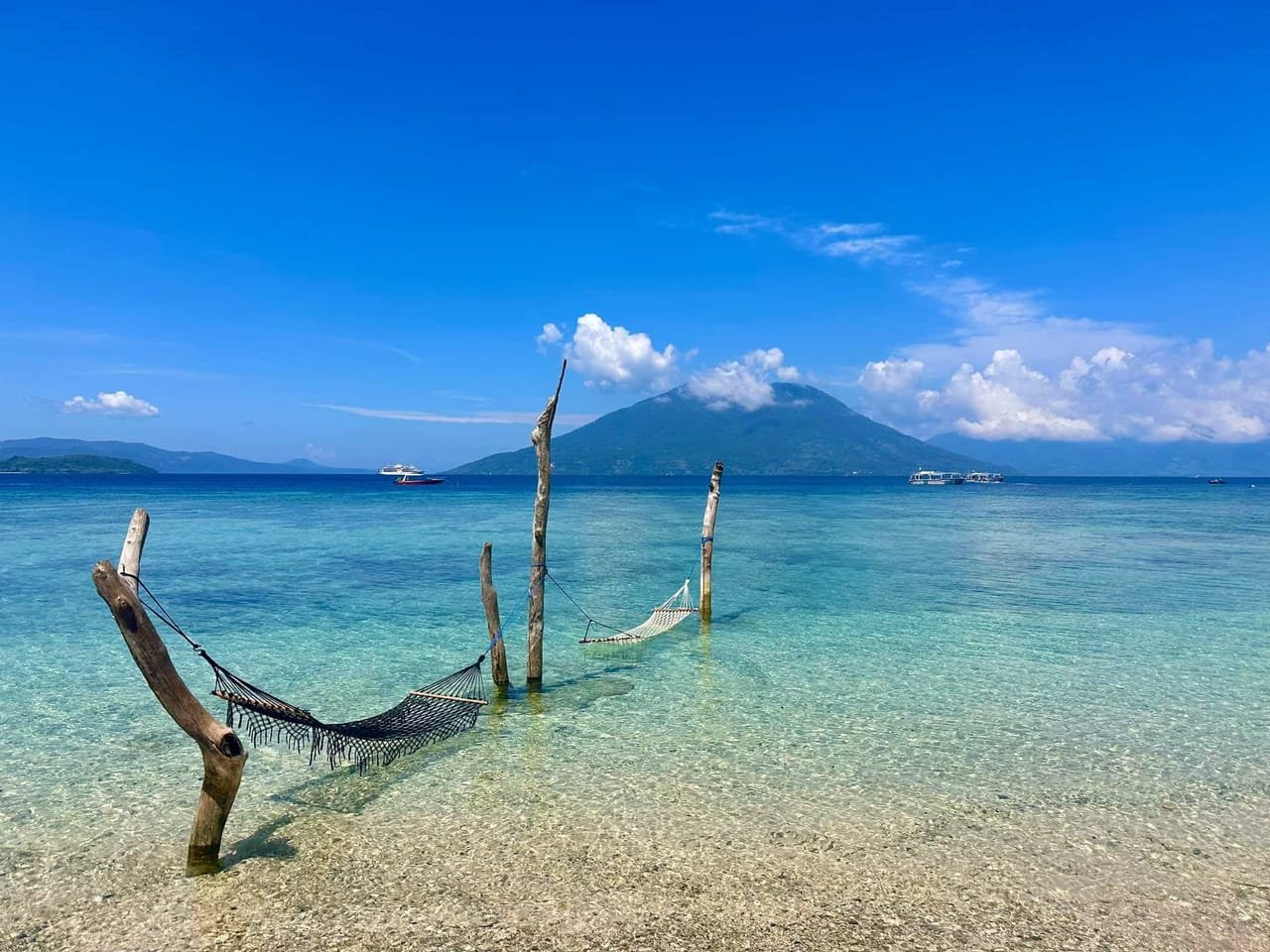 The house reef beach with hammocks in shallow turquoise water and the Pantar volcano in the distance