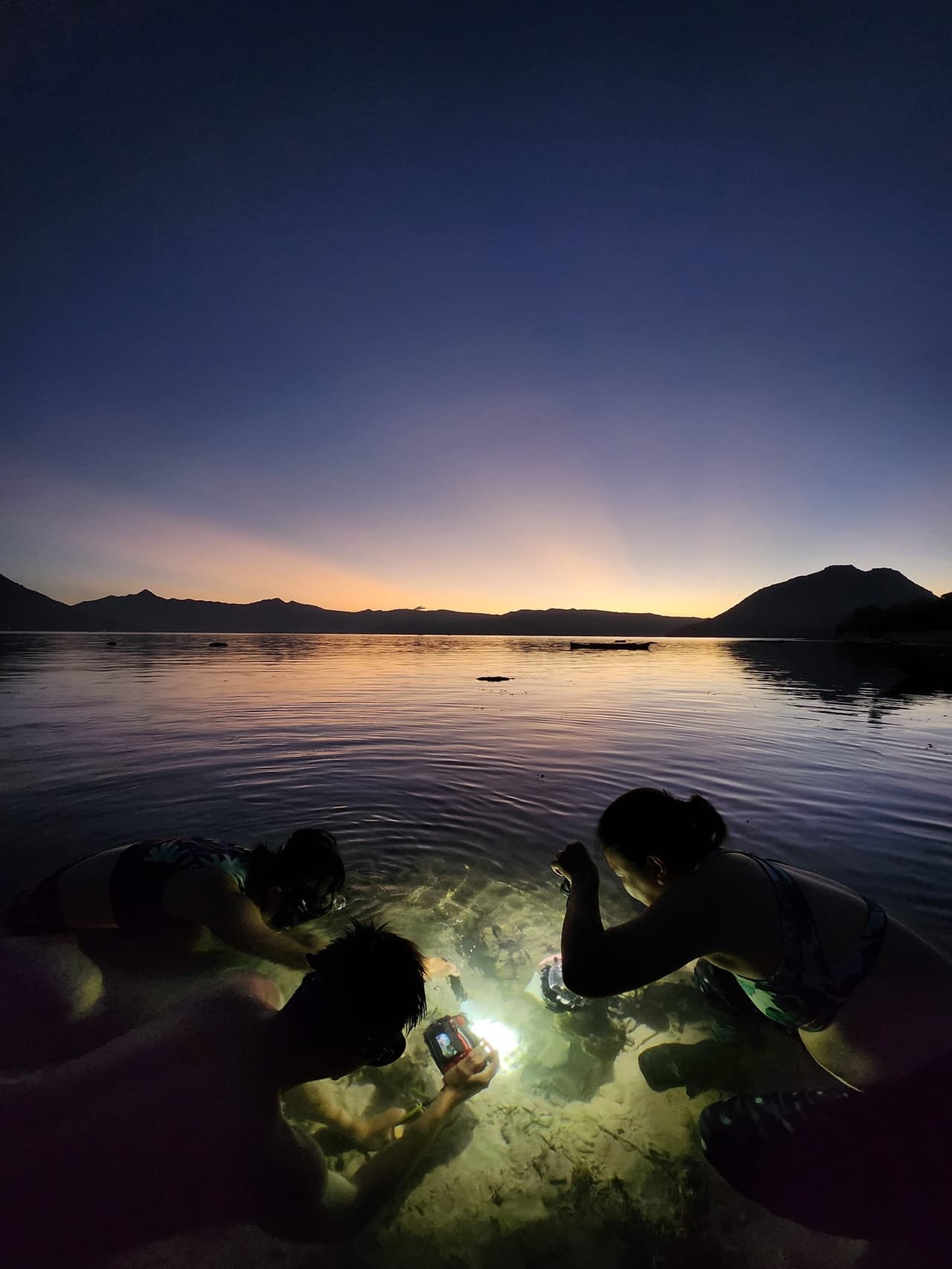 Two divers searching a tide pool at sunset with a torch, Pantar Strait in the background